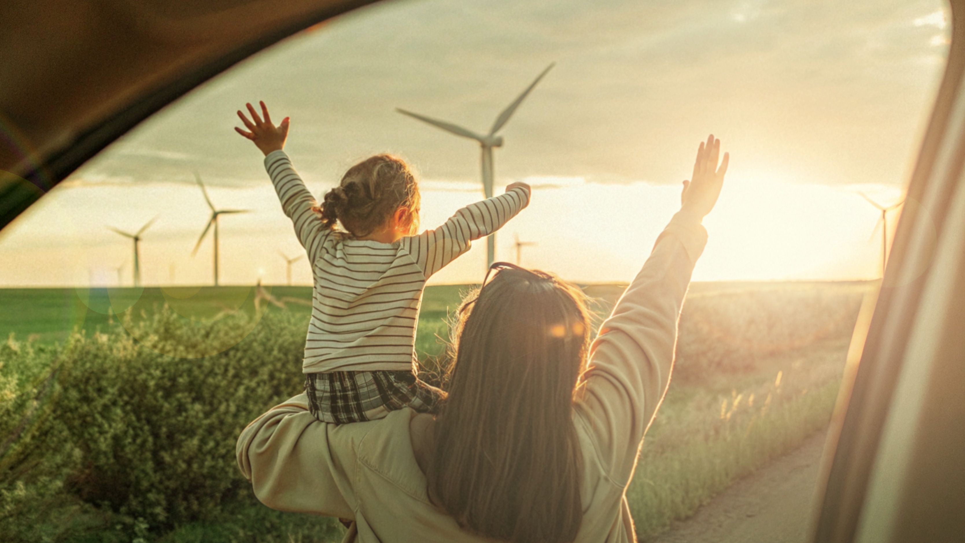 Una donna con sua figlia in spalla guardano, serene, verso l'orizzonte nel quale ci sono delle pale eoliche